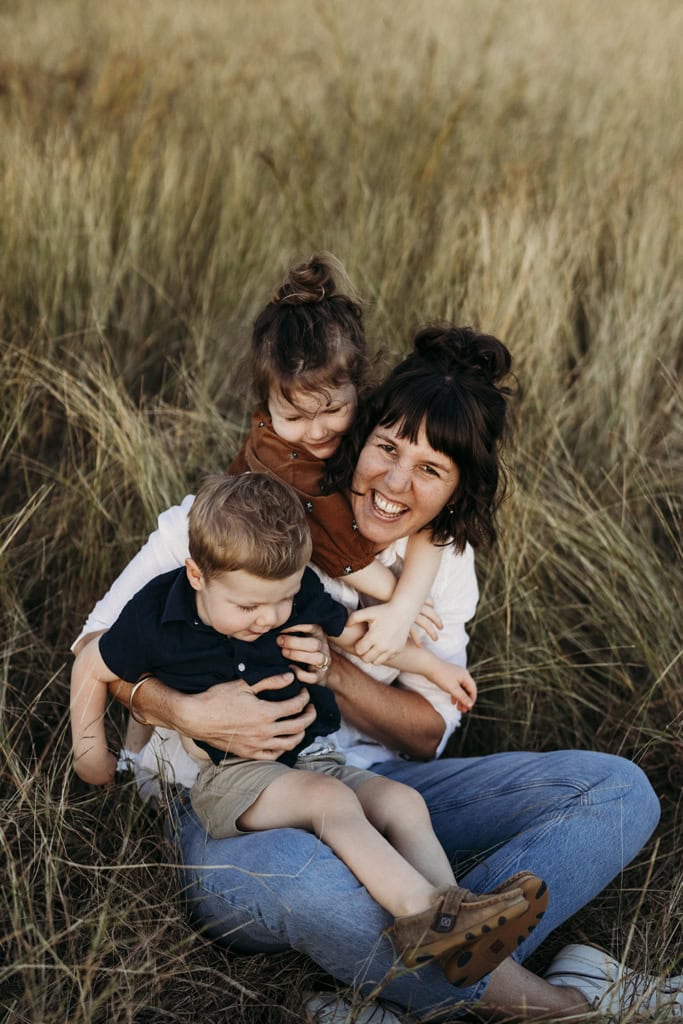 A mother sits in long grass laughing as she cradles her two small children.