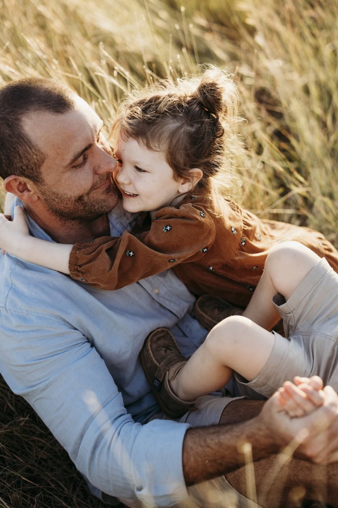 A father sits in long grass and cradles his daughter. His son's legs are visible as the father holds his hands but the little boy's face is out of shot. 