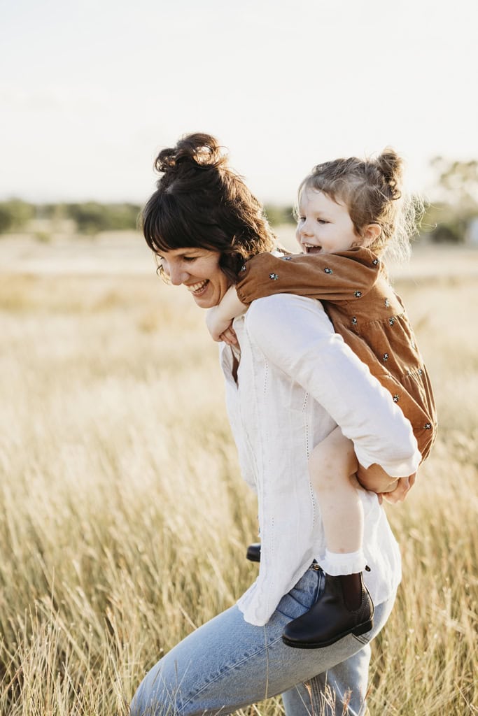 A mother gives her young daughter a piggy back as she runs through long grass.