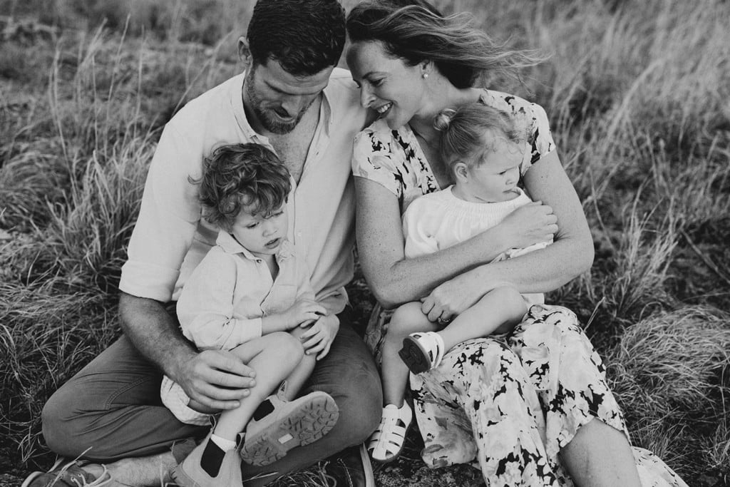 A family of four sit on the ground and cuddle as they laugh.