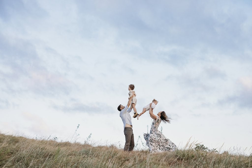 A man and woman hold their children above their heads playfully as their children laugh. They are standing on top of a grassy hill as the wind blows their hair. 