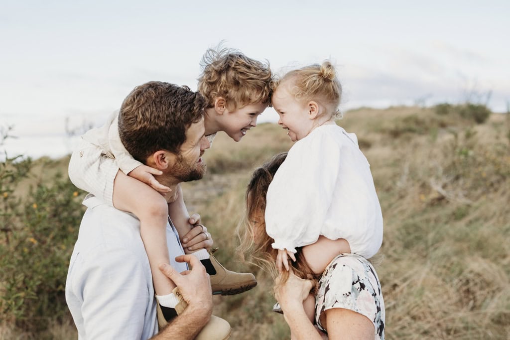 Two small children sit on top of their parents' heads and lean towards each other and laugh.