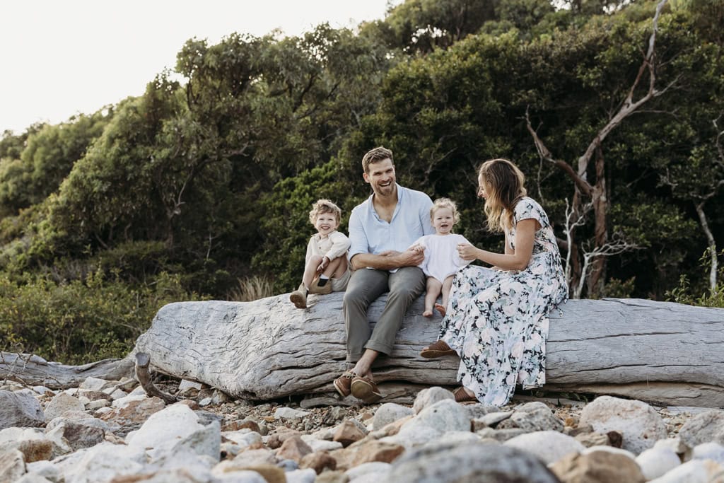 A family of four sit on a large log at the beach. 