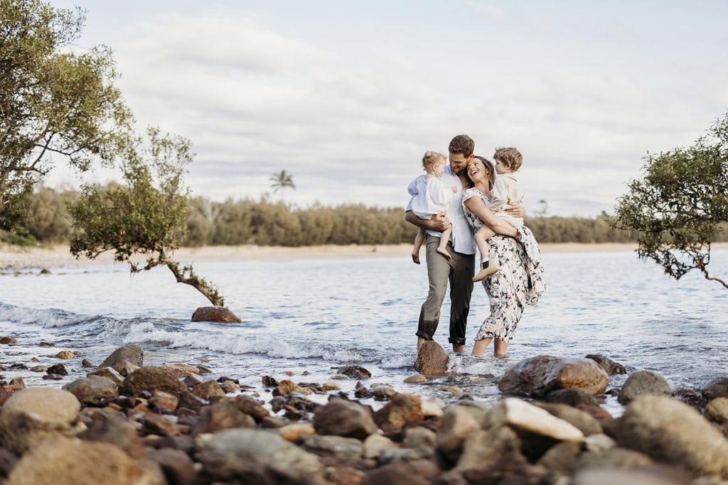 A family of four stand on a shallow beach in the water. Rocks surround them as they cuddle one another. 