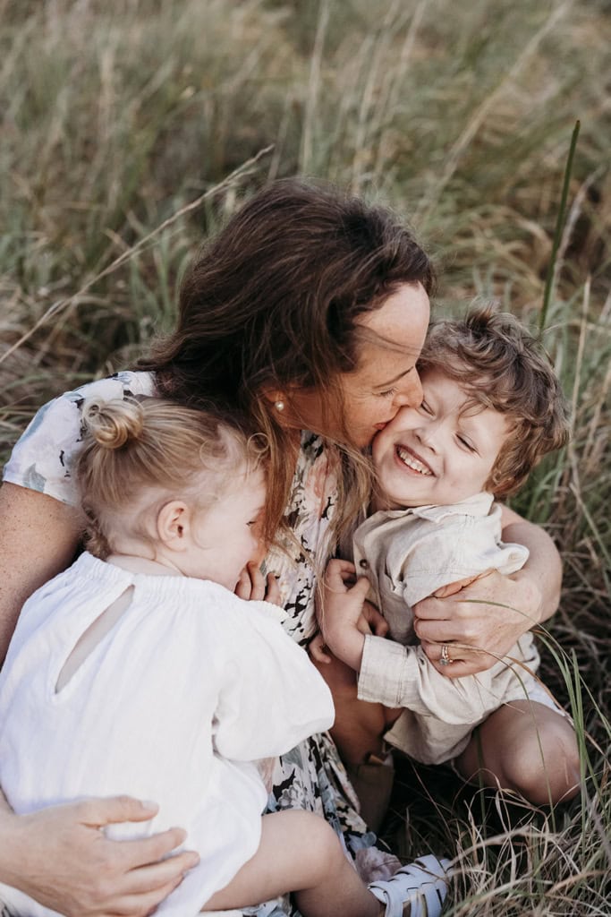 A mother sits in long grass with her two children, as she kisses her son's cheek. He is smiling. 