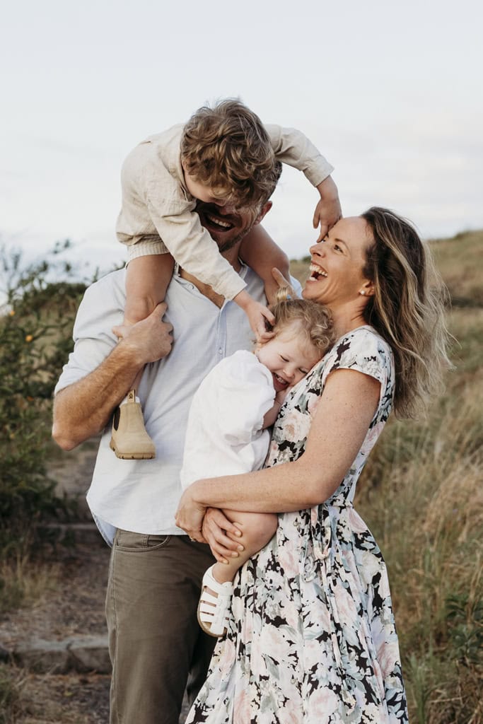 A family of four laugh at one another. A mother holds her toddler to her chest, while the father has his son on his shoulders. The little boy leans down and tickles his little sister. 