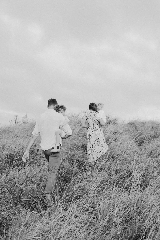 A couple walk up a grassy hill as they hold their two small children in their arms. 