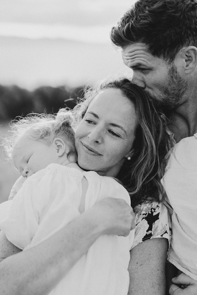 A man kisses his wife on top of her head as they snuggles their daughter. 