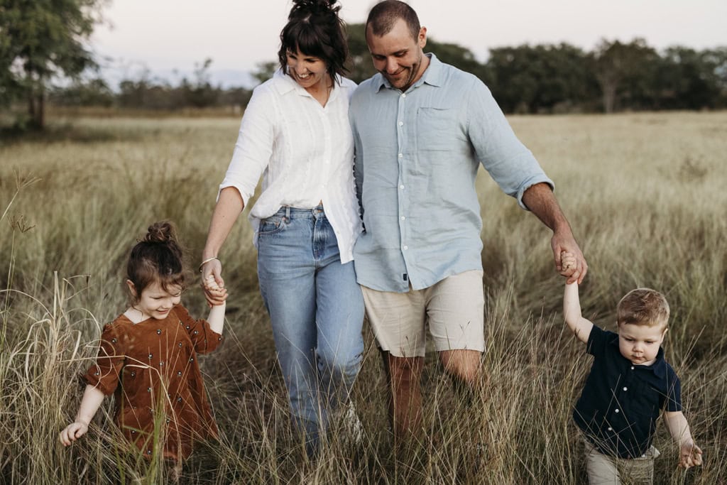 A family of four hold hands as they walk through long grass. The couple's young children lead them. 