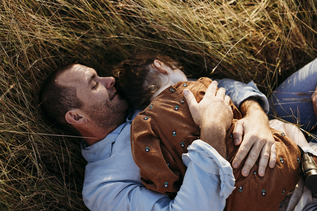 A father lies in long grass as he cuddles his daughter to his chest.