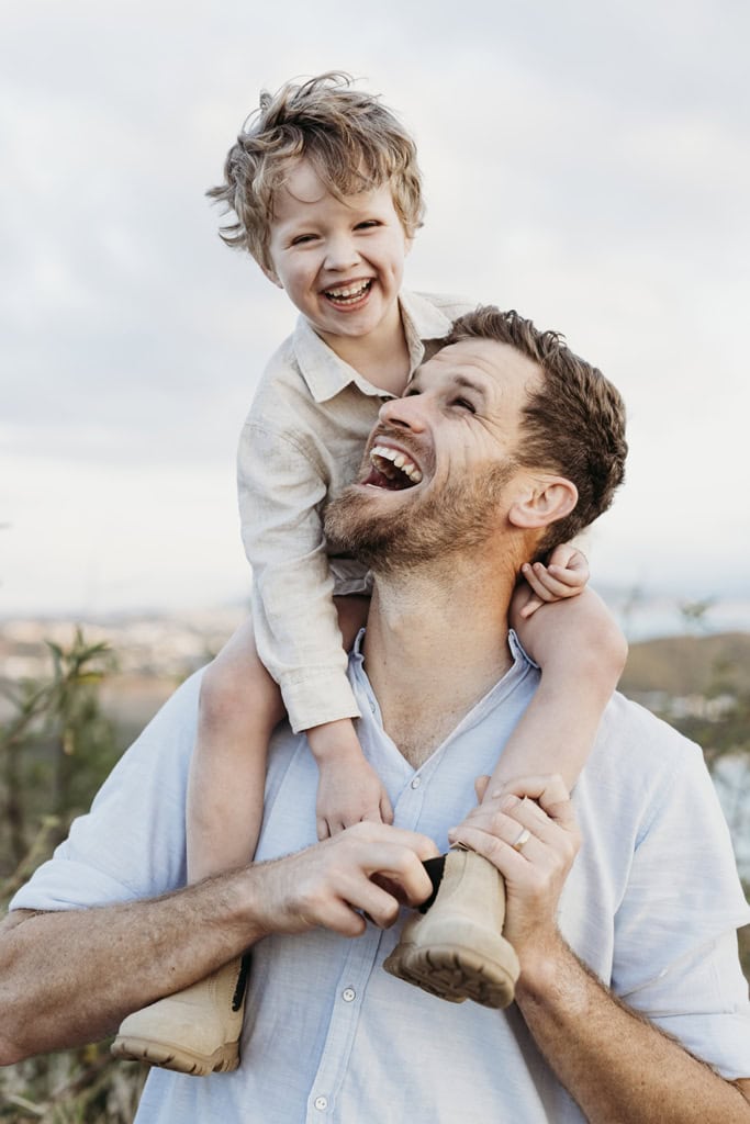 A man looks up at his son who is on his shoulders, as they both laugh. 