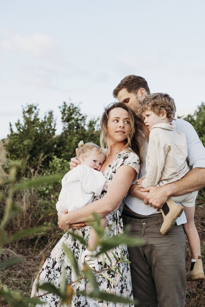 A family of four snuggle together, standing in bushland.