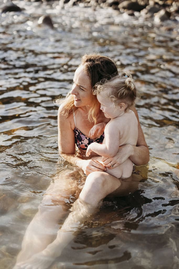 A mother sits in water with her toddler.