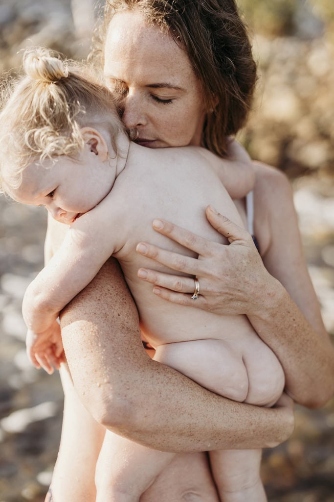 A mother holds her toddler daughter against her chest and breaths her in at the nape of her neck. 