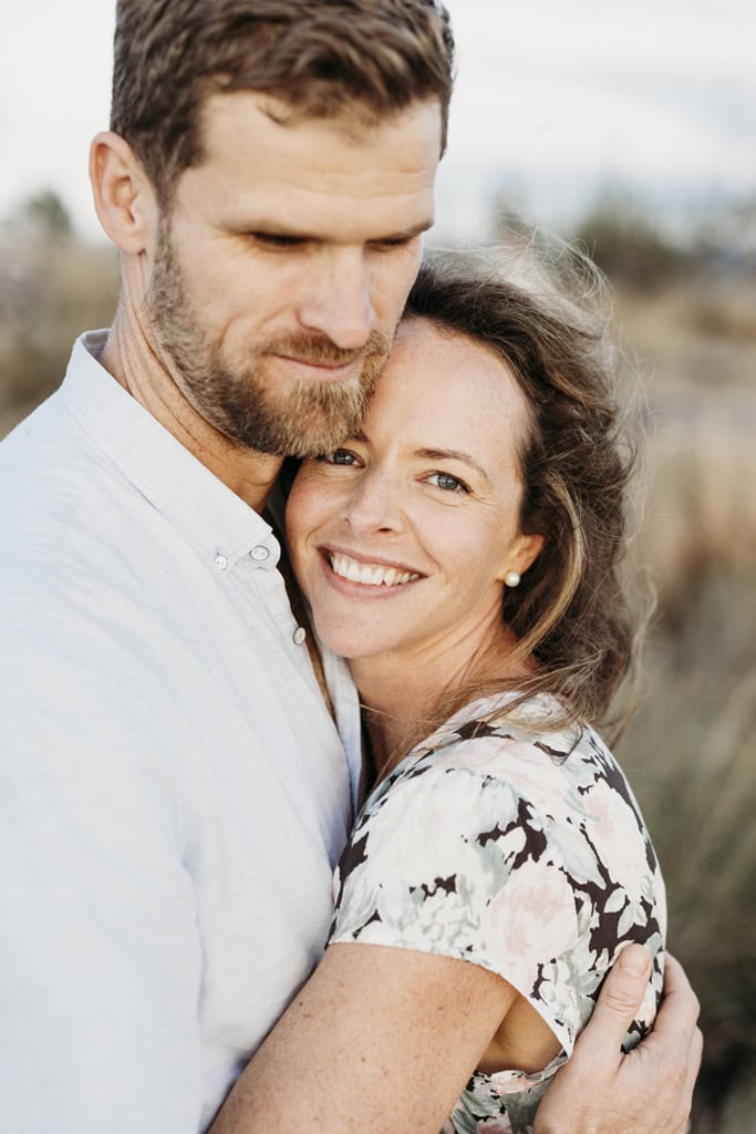 A woman hugs her partner and has her face pressed against his chest. She is looking at the camera smiling.