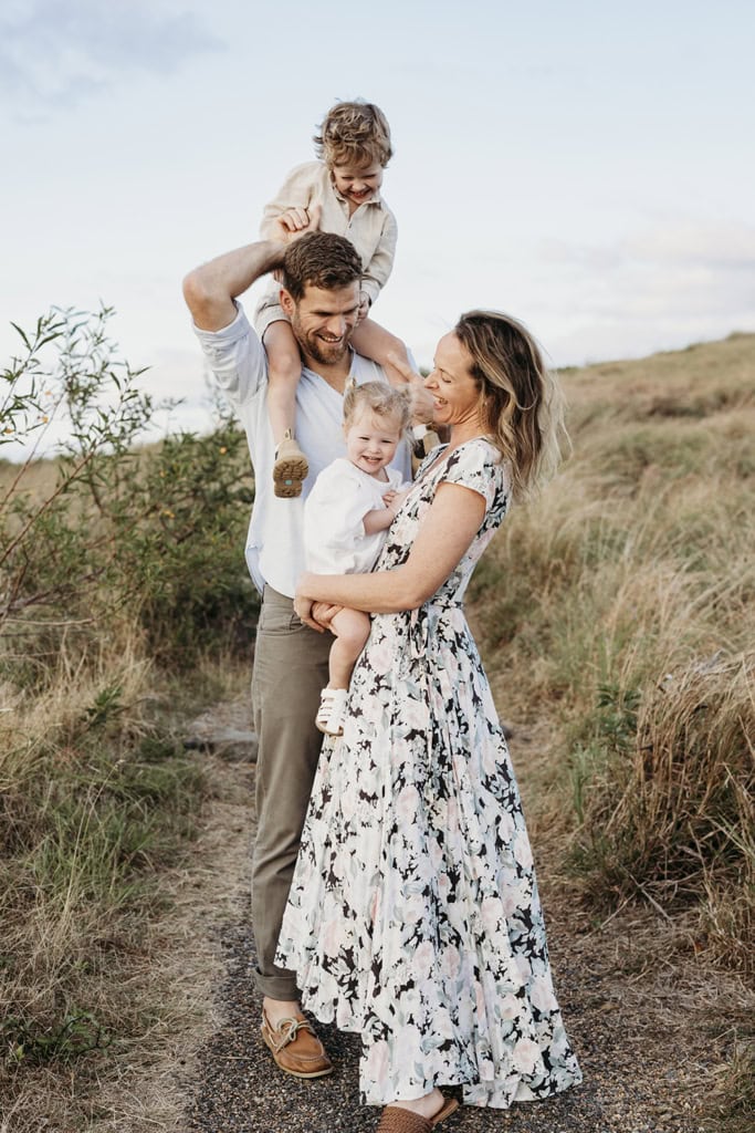 A family of four laugh. A father has his son on his shoulders, while a woman holds her daughter in front of her.